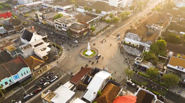 Aerial drone footage of Tugu Yogyakarta at a major urban intersection, surrounded by city traffic, roads, buildings, and warm morning sunlight in Yogyakarta, Indonesia.