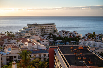 The sun drops toward the Atlantic behind the curved white hotel and terracotta rooftops of Los Gigantes. A band of pale gold spreads across the water and the sky. © Oksana