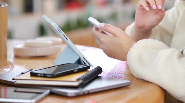 Person uses smartphone while tablet and phone sit nearby on wooden table. Hands hold device close to eye. Warm lighting, cozy indoor setting visible. Multiple tech devices arranged on surface