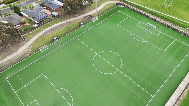 Aerial top-down view of a modern synthetic turf soccer pitch with marked field lines, adjacent to residential homes and sports fields in suburban Melbourne, Victoria, Australia. Community facility