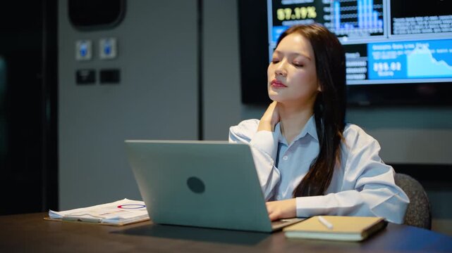 Asian businesswoman working late at night in modern office feels exhausted and suffers from neck pain while typing on laptop computer, dealing with severe office syndrome and workplace stress.