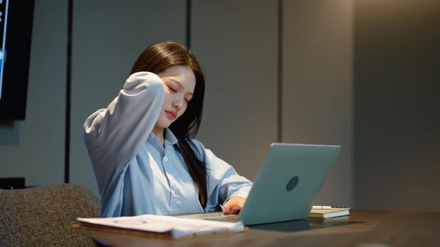 Asian businesswoman working late at night in modern office feels exhausted and suffers from neck pain while typing on laptop computer, dealing with severe office syndrome and workplace stress.