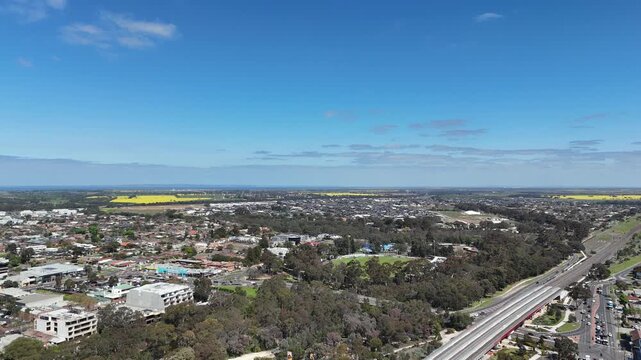 Aerial drone view of Werribee township with commercial buildings, residential housing, railway tracks, road traffic, the tree-lined Werribee River and distant golden canola fields under a blue sky. 