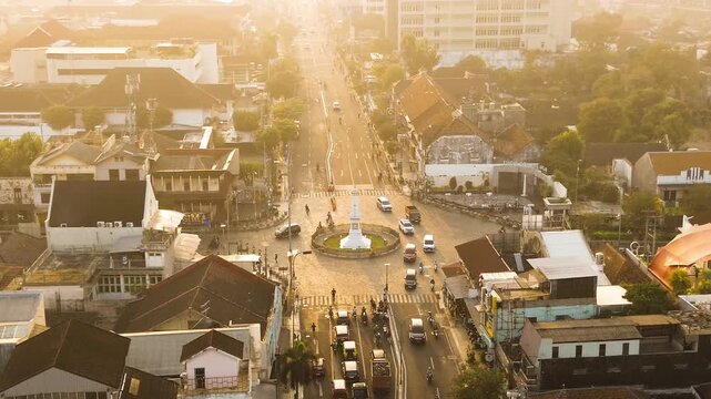 Aerial drone footage of Tugu Yogyakarta at a busy city intersection, surrounded by traffic, urban buildings, and warm golden morning sunlight in Yogyakarta, Indonesia.
