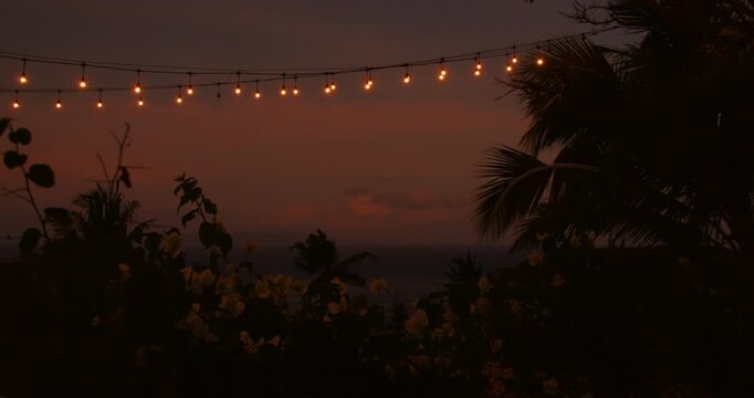 Warm string lights glow against pink sunset sky above the tropical ocean