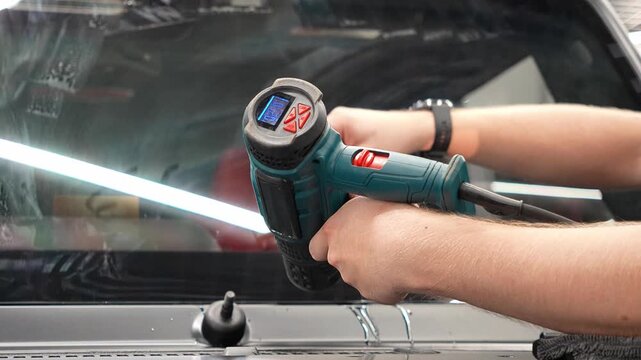 In the process of installing transparent polyurethane film on a car windshield to prevent stone chips, warming up the wrap with a gun and positioning it is carried out. Professional auto detailer. 
