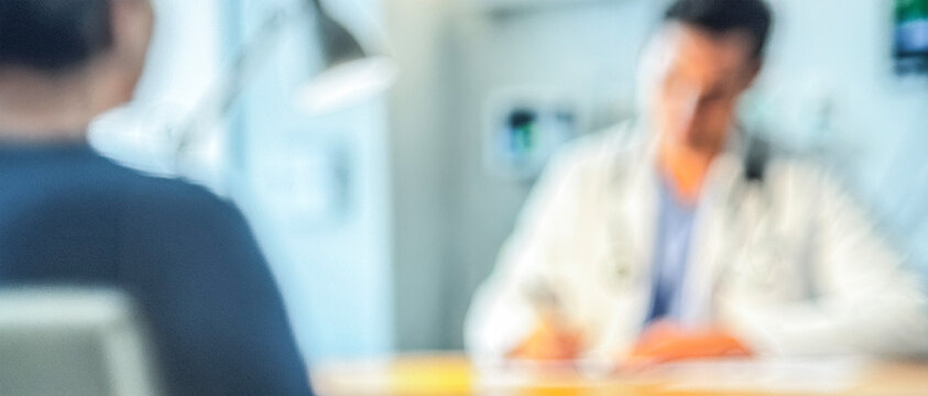 A healthcare professional is seen from a blurred perspective while engaging with a patient in a clinical setting, emphasizing a confidential discussion.
