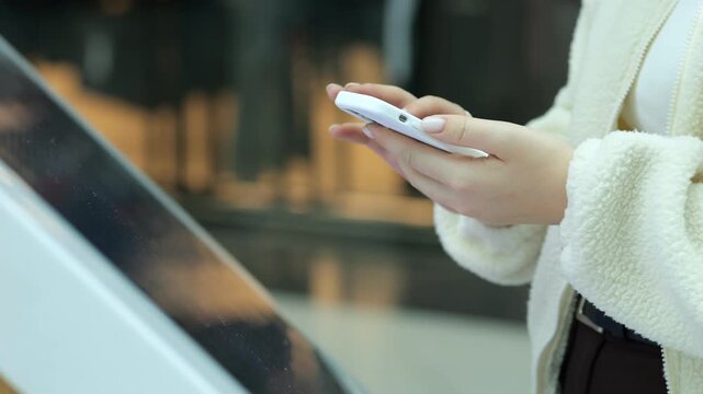 Person holds phone while touching interactive screen. Wears light-colored jacket and dark pants. Background shows blurred indoor setting. Focused on interaction with digital display
