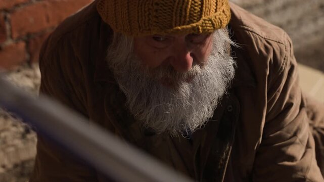 Senior Caucasian homeless man sitting outdoors while camera tilts up from hands holding coin. Clip ideal for charity campaigns, social awareness projects and documentary editing