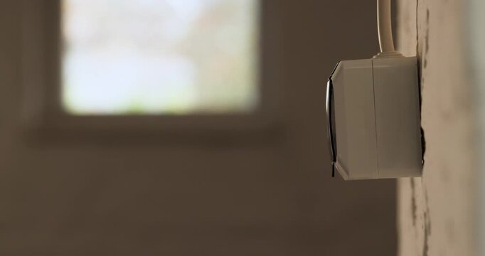 Adult hand plugs a modern smartphone charger into a wall power outlet inside a dimly lit garage room.
