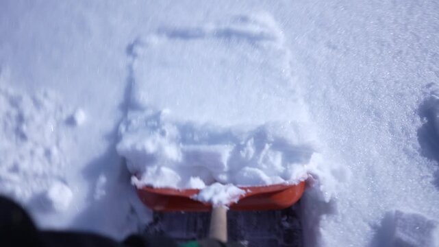 Point of view of snow removal with an orange shovel clearing fresh snow from a walkway on a sunny winter day outdoor seasonal maintenance concept, close up, slow motion