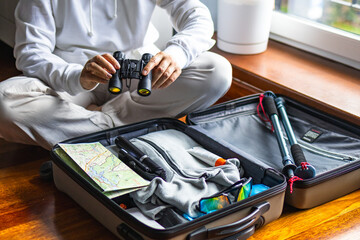Woman sits cross legged holding binoculars before placing them into suitcase full of hiking gear. Concept of navigation equipment preparation and trip packing checklist for mountain hiking trip. © Ina