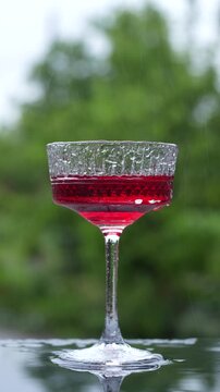 Red cocktail in textured coupe glass on dark reflective surface, outdoors, during the rain. Close up of refreshing red drink or cocktail in crystal coupe glass against blurred green nature background