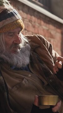 Vertical video of senior Caucasian homeless man counting collected coins in metal tin on city street. Clip suitable for charity campaigns and poverty awareness videos