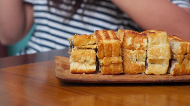 Front angle video of a woman taking a piece of toast bread from a wooden plate beside a glass of coffee. Cozy breakfast scene.
