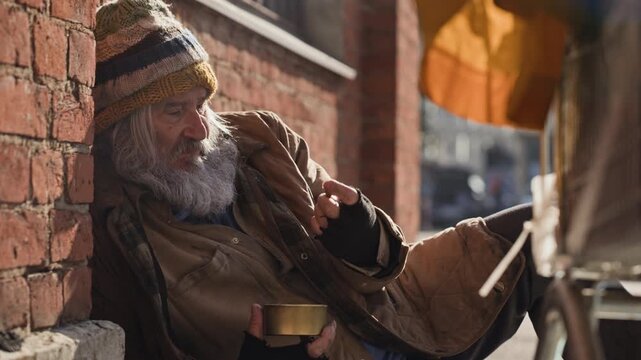 Senior Caucasian homeless man sitting on city sidewalk and counting coins in metal tin. Perfect for poverty awareness campaigns, charity documentaries, and social aid projects