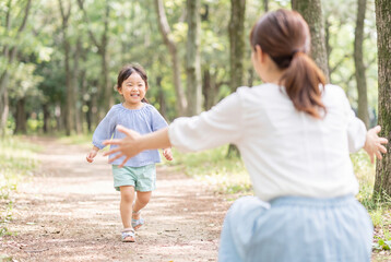 Obraz premium Maman vue de dos, accroupie, bras ouverts, en chemise blanche et jupe bleu clair, accueillant sa petite fille asiatique souriante courant en haut rayé bleu et blanc, short vert pastel et sandales