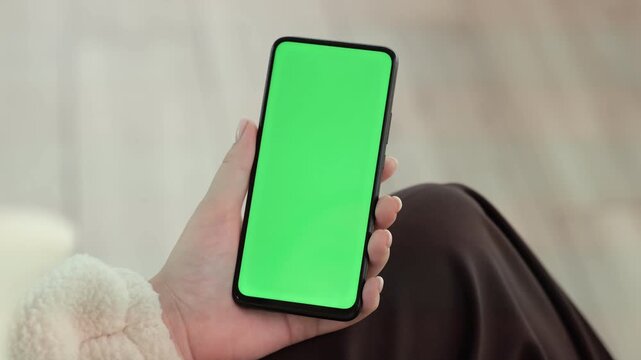 Hand holds phone with green screen. Phone screen shows solid green. Background blurred with soft light. Person wears cozy sweater. Legs rest on chair. Phone held steady for easy use