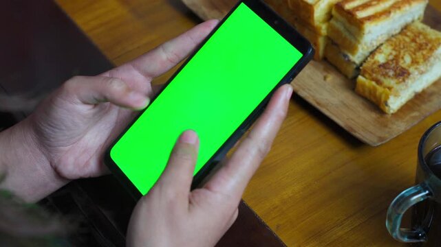 Close-up video of hands using a green screen phone on a wooden table beside toast and coffee. Perfect mock-up template for apps.
