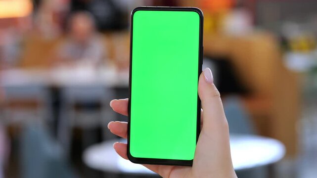 Hand holds smartphone with green screen. Background blurred cafe setting. Phone held steady, screen fully green. Urban cafe ambiance with tables and chairs. Focused on phone and hand interaction