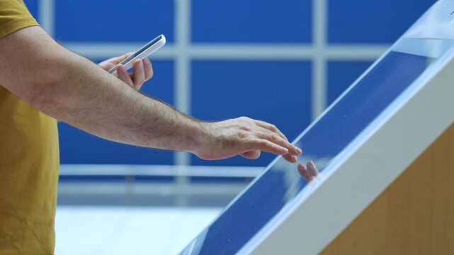 Man interacts with touchscreen display while holding smartphone. He taps and swipes on glossy surface. Background features blue windowed wall. Scene shows modern tech usage in public space