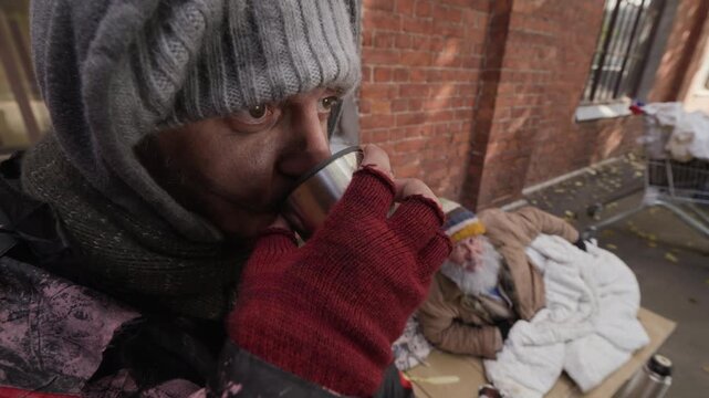 Caucasian young adult homeless man drinking hot beverage and eating bread near senior Caucasian companion on sidewalk. Useful for social welfare and charity campaigns