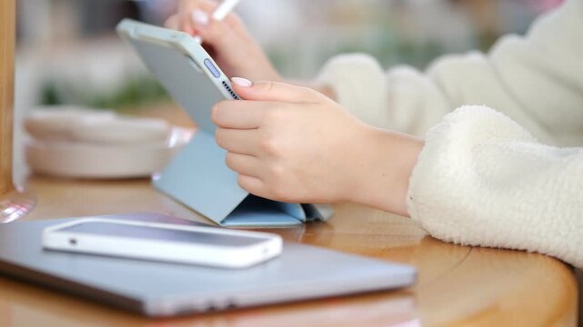 Person uses stylus on tablet at wooden table. Phone and laptop sit beside tablet. Hands wear white knitted sweater. Warm indoor lighting. Blurred background with natural tones