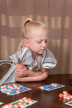 Young blonde girl sits at a wooden table playing a card and token game, concentrating thoughtfully while developing cognitive and strategic skills during quiet indoor playtime