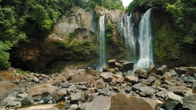 Woman in swimsuit walks across rocks beneath tall tropical waterfall in jungle by dense forest