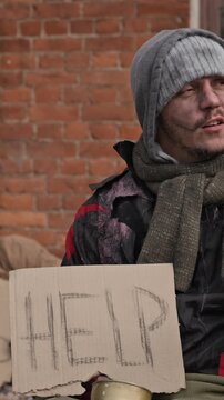 Vertical shot of homeless Caucasian young adult man sitting on street holding cardboard sign asking for help. Useful for charity promotions and social welfare documentaries