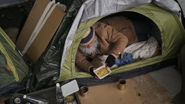 Senior Caucasian homeless man lying inside street tent eating noodles from container. Perfect for charity campaigns, poverty awareness videos, and humanitarian aid reports