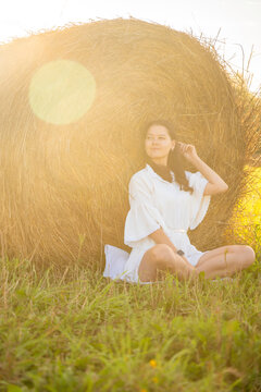 Young woman in white dress relaxing beside a hay bale in warm sunset light. Peaceful countryside moment represents slow living, solitude and connection with nature