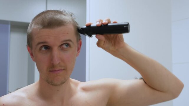 Bare chested man uses cordless clippers for self haircut beside bathroom mirror. He works around short hair with steady motions, creating even buzz cut and showing quiet concentration throughout.