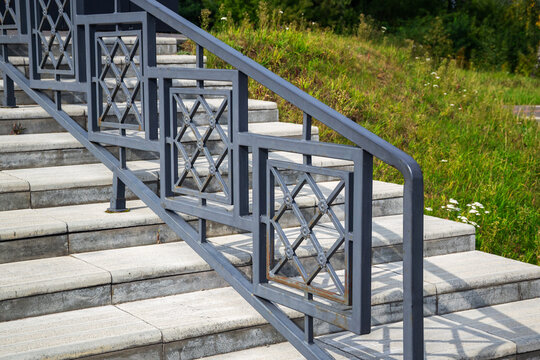 Ornate metal railing on outdoor concrete stairs near green grassy slope on sunny day