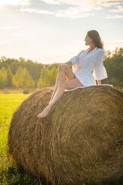 Young woman in white dress sitting barefoot on hay bale in summer countryside. Peaceful rural lifestyle represents slow living, freedom and relaxation in nature