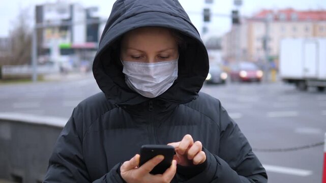 Woman in face mask texts on smartphone beside urban roadway. Winter hood frames her face as she remains still and intent, with passing traffic and apartment blocks creating cool city mood.