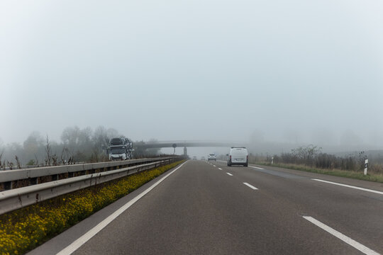 Fog covers German motorway during autumn morning, reducing visibility around cars, van, truck and overpass. Misty highway scene shows wet roadside, traffic lanes and challenging driving conditions