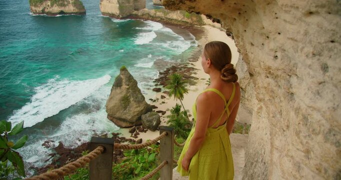 Young woman tourist admires coastal landscape from cliff stairs at Diamond Beach in Bali