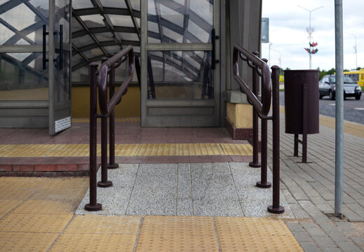 Accessible entrance ramp with brown metal handrails and yellow tactile paving tiles at public transit station
