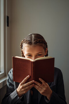 Vertical composition of young schoolgirl with braids reading book obscuring hands in natural window light in calm neutral mood generative AI