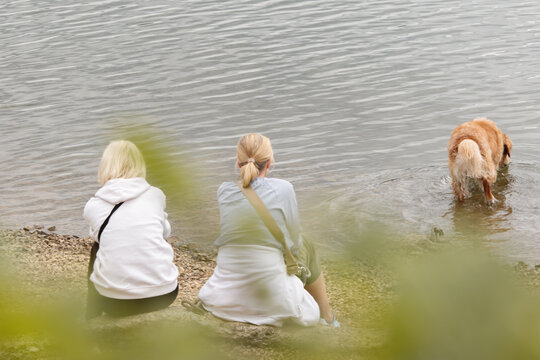 Zwei Frauen sitzen am Ufer. Der Hund spielt im Wasser.