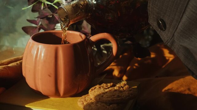 Autumn vibes. Pouring tea into mug from teapot. Cup of hot drink on windowsill in cozy interior. Cozy fall mood. 