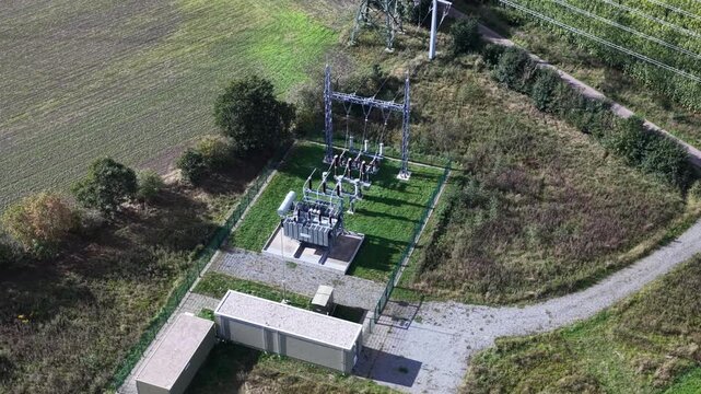 Aerial footage of compact electrical substation feeding renewable wind or solar energy into the power grid, located in a rural field next to a transmission tower.