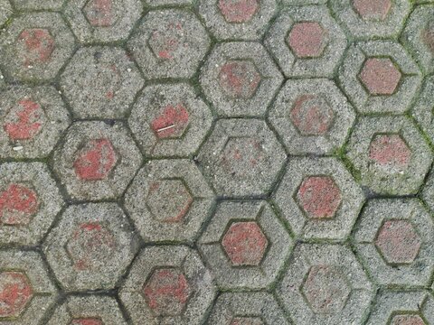 Top view of weathered hexagonal concrete paving blocks with faded red centers and green moss growing in the joints. A geometric texture