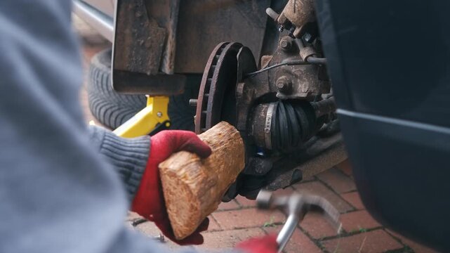 Mechanic striking wooden block to free seized brake rotor from wheel hub. Automotive repair removing stuck brake disc during car maintenance with manual tools. Closeup disassembly process with