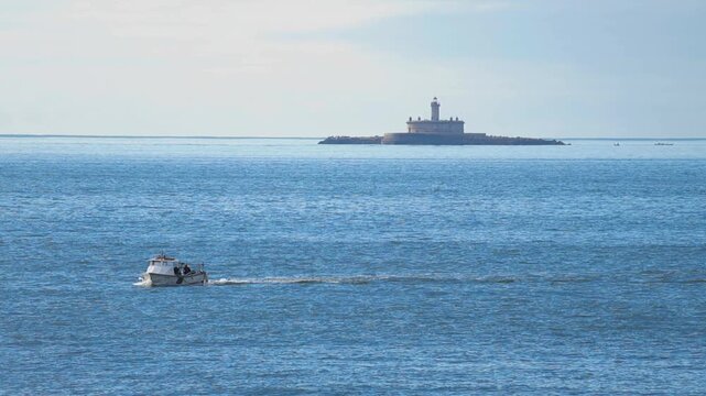 Distant view of a small motorboat sailing on the deep blue waters of the Tagus river estuary with the historic Bugio Lighthouse visible on the horizon under a hazy sky near Lisbon, Portugal