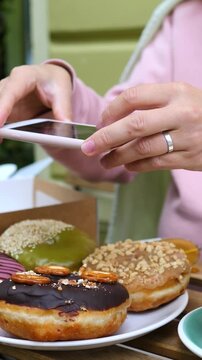 Hands frame smartphone above donuts while photographing dessert on table. Pink sleeve and silver ring stand out as fingers steady device near chocolate, sesame, and green glazed pastries.