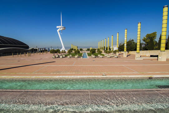 View of the Montjuic Communications Tower rising above the plaza with its white structure, yellow pillars, and a water feature in Barcelona, Catalonia, Spain.