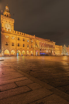 View of the historic Palazzo d'Accursio with its clock tower illuminated at night, featuring arched porticoes and a festive Christmas tree in Bologna, Emilia-Romagna, Italy.
