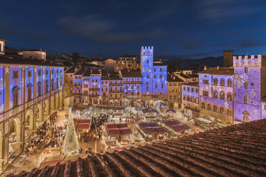 View of the Christmas market in Piazza Grande with festive light projections on historic buildings and a large Christmas tree in Arezzo, Tuscany, Italy.